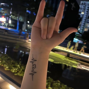 May include: A black and white tattoo of a heartbeat with a heart in the middle on a person's wrist.