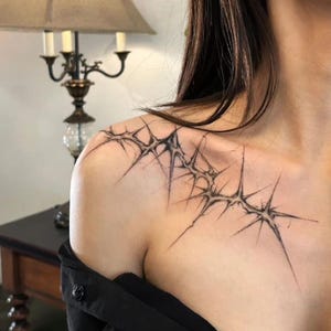 Op de afbeelding: Een zwart tattoo-ontwerp op de schouder en het sleutelbeen. De tattoo heeft een stekelig, stervormig patroon met fijne lijnen en schaduwen, wat een gedetailleerde en artistieke uitstraling creëert. Het ontwerp is gecentreerd op de schouder en loopt over het sleutelbeen.
