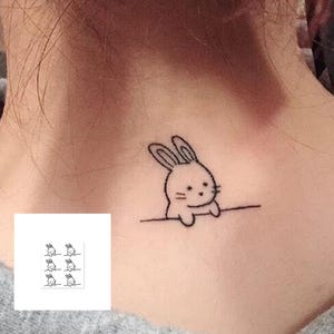 May include: A small, black outline tattoo of a cartoon bunny peeking over a line is visible on the skin. The bunny has large ears and a cute face. A sheet of six similar bunny temporary tattoos is in the lower left corner.