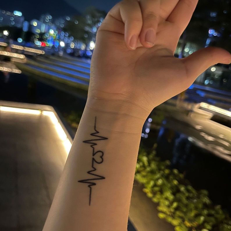 May include: A black ink tattoo of a heartbeat with a heart in the middle on a person's wrist.