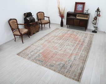 May include: A rectangular, distressed-look rug with a faded, vintage aesthetic. The rug features a central field of muted coral and beige tones, with accents of blue and brown. The rug is placed on a light-colored wooden floor, with antique furniture in the background.