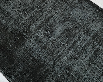 May include: A rectangular, distressed black rug with a textured surface. The rug has a faded appearance with lighter areas, creating a vintage or antique aesthetic. The rug is placed on a white surface.