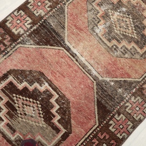 May include: A distressed, rectangular rug with a geometric pattern. The rug features a central design in shades of pink and brown, framed by a darker brown border with repeating geometric shapes. The rug has a vintage, worn appearance.