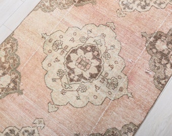 May include: A vintage rug with a faded pink background and a central medallion in shades of beige, brown, and pink. The rug has a distressed look with frayed edges and a worn appearance.