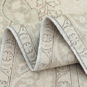 Pode incluir: Close-up de um tapete dobrado, em estilo vintage, com um aspeto desbotado e desgastado. O tapete apresenta uma paleta de cores bege e azul claro com intrincados padrões florais e geométricos. As bordas são delimitadas com uma borda castanha mais escura.