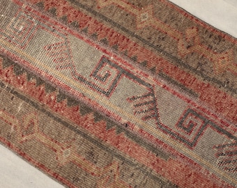 May include: Close-up of a distressed, rectangular rug with a faded, geometric pattern. The rug features a color palette of muted reds, grays, and browns. The design includes repeating geometric shapes and lines, creating a vintage aesthetic.
