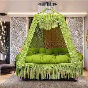 Op de afbeelding: Een groene hangende hangmatstoel met een kap en franjes. De stoel is gemaakt van macramé en heeft een dik, comfortabel kussen.