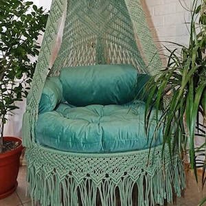 May include: A hanging macrame chair in a soft green hue. The chair features a round, plush seat and back cushions in a matching color. The macrame design includes intricate knotting and fringe details, creating a bohemian aesthetic. The chair is suspended from a ceiling hook.