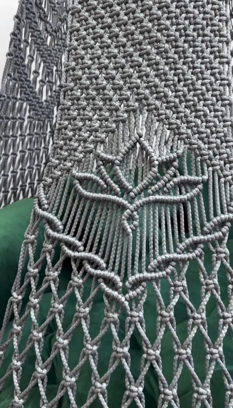 May include: Close-up of a gray macrame textile with a leaf-like design. The handmade piece features intricate knotting patterns and a diamond-shaped net. The background is a dark green fabric.