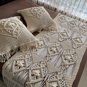 May include: A cream-colored macrame throw blanket with a geometric pattern and fringe. Two matching macrame pillows are on the bed behind the blanket.