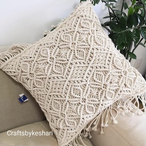 Op de afbeelding: Een crèmekleurig macramé kussen met een geometrisch patroon en franjes. Het kussen is gemaakt van katoenen koord en heeft een vierkante vorm.