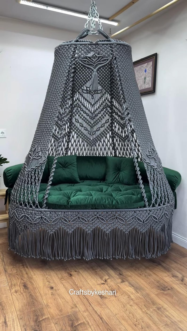 May include: A gray macrame swing bed with a canopy and fringe. The bed features a green velvet cushion and pillows. The canopy has a decorative pattern. The bed is suspended from the ceiling.