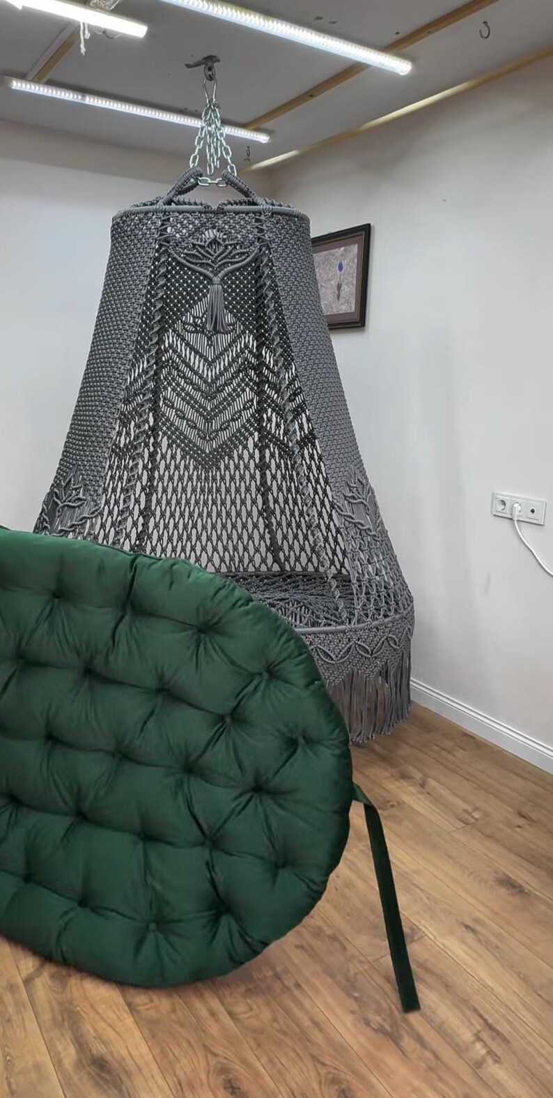 May include: A gray macrame hanging chair with a decorative fringe and a dark green tufted cushion. The chair is suspended from the ceiling by a chain. The cushion is round and has a strap.