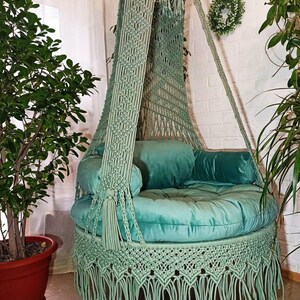 May include: A mint green macrame hanging chair with a round, cushioned seat and back pillows. The chair features a fringed bottom and is suspended from a wooden beam. Decorative tassels and macrame detailing add to the bohemian style.