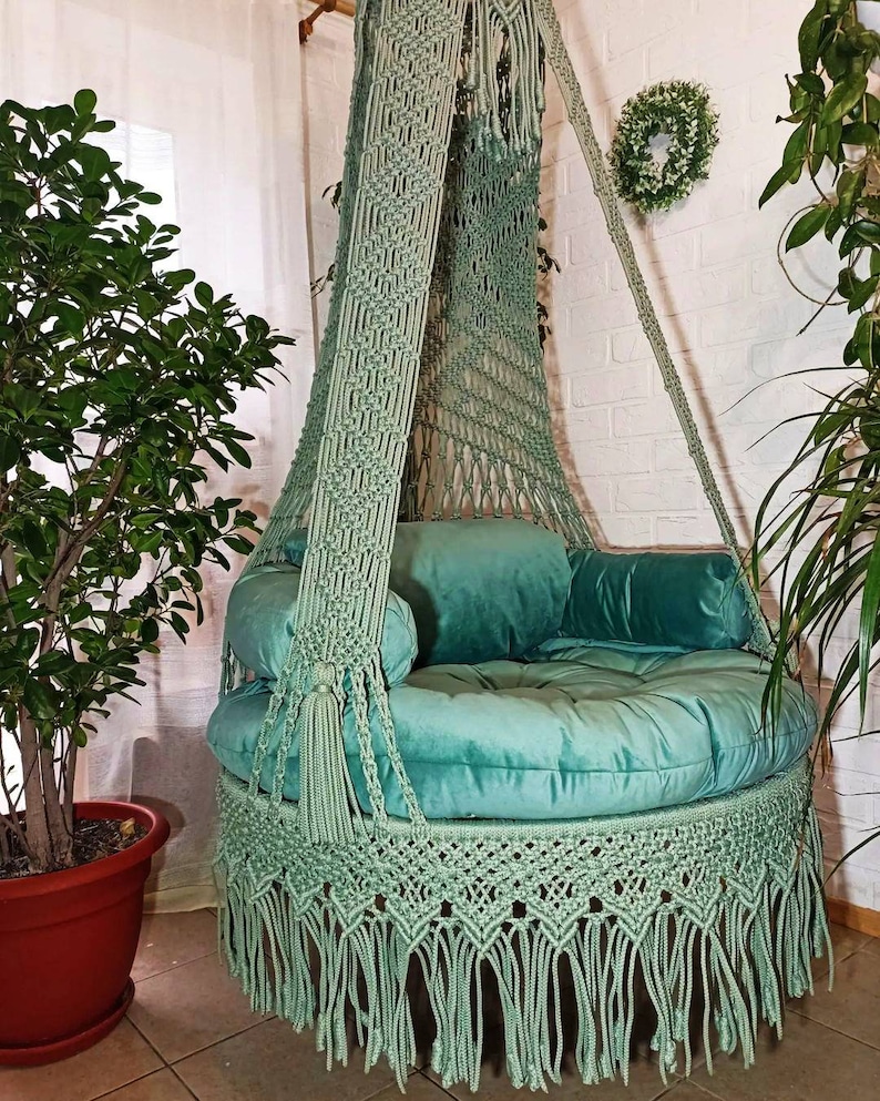 May include: A mint green macrame hanging chair with a round, cushioned seat and back pillows. The chair features a fringed bottom and is suspended from a wooden beam. Decorative tassels and macrame detailing add to the bohemian style.