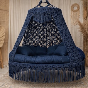 May include: A navy blue macrame hanging daybed with a fringed base and canopy. The round bed has a cushioned base and pillows. The background includes woven textures, plants, and a woven vase, creating a bohemian aesthetic.