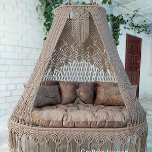 May include: A handcrafted macrame swing bed in shades of brown, featuring intricate knotwork and a fringed edge. The bed includes plush brown cushions and pillows, creating a cozy and stylish seating area. The swing is suspended from a metal chain.