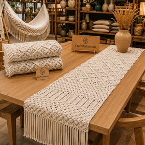 May include: An off-white macrame table runner with a geometric pattern and long fringe ends, displayed on a wooden table alongside a folded macrame runner and a small wooden sign featuring the text Craftsbykeshari in a clear, legible font.