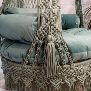 May include: A sage green macrame hanging chair with a round seat and backrest. The chair features a woven design with a decorative fringe along the bottom edge. Soft, plush cushions in a matching green color provide comfort. A tassel hangs from the front.