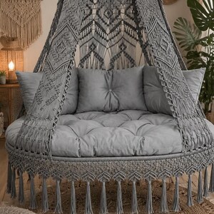 Peut inclure: Un lit suspendu en macramé gris avec une base de coussin ronde et touffetée et des oreillers assortis. Le lit est orné d'un auvent décoratif et de franges, créant une esthétique bohème. Le lit est posé sur un tapis en jute tissé.