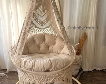 May include: A beige macrame hanging chair with a fringe trim and a thick beige cushion. The chair is hanging from a wooden frame and is in a room with white curtains.