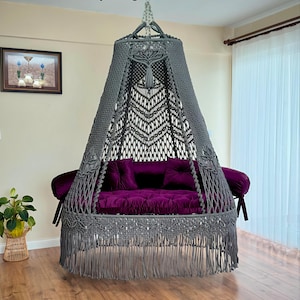 Peut inclure: Un fauteuil suspendu en macramé gris avec un coussin en velours violet et une bordure en franges. Le fauteuil est suspendu au plafond et possède un toit en forme de canopée.