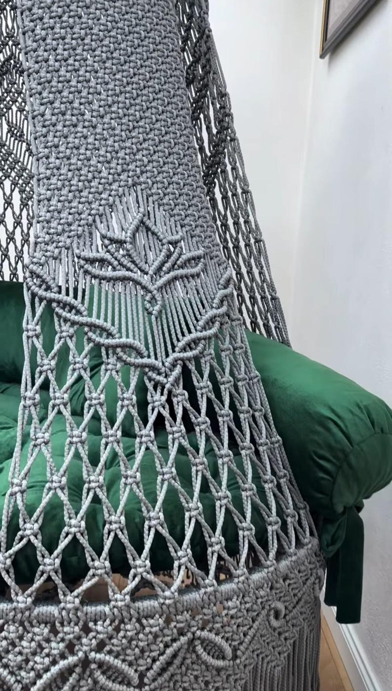 May include: A gray macrame hanging chair with a green cushion and pillow. The chair features intricate knotting patterns and a decorative leaf design. The chair is suspended from the ceiling, creating a cozy and stylish seating area.