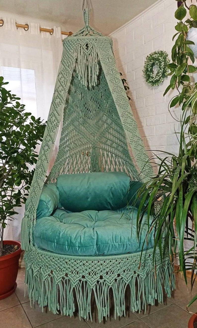 May include: A hanging macrame chair in a soft green hue. The chair features a round, plush seat and back cushions in a matching color. The macrame design includes intricate knotting and fringe details, creating a bohemian aesthetic. The chair is suspended from a ceiling hook.