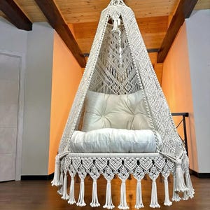 Peut inclure: Un fauteuil suspendu en macramé crème avec un coussin et un oreiller blancs. La chaise est suspendue au plafond et présente des motifs tissés complexes et des franges décoratives. Un ajout confortable et élégant à tout espace intérieur.