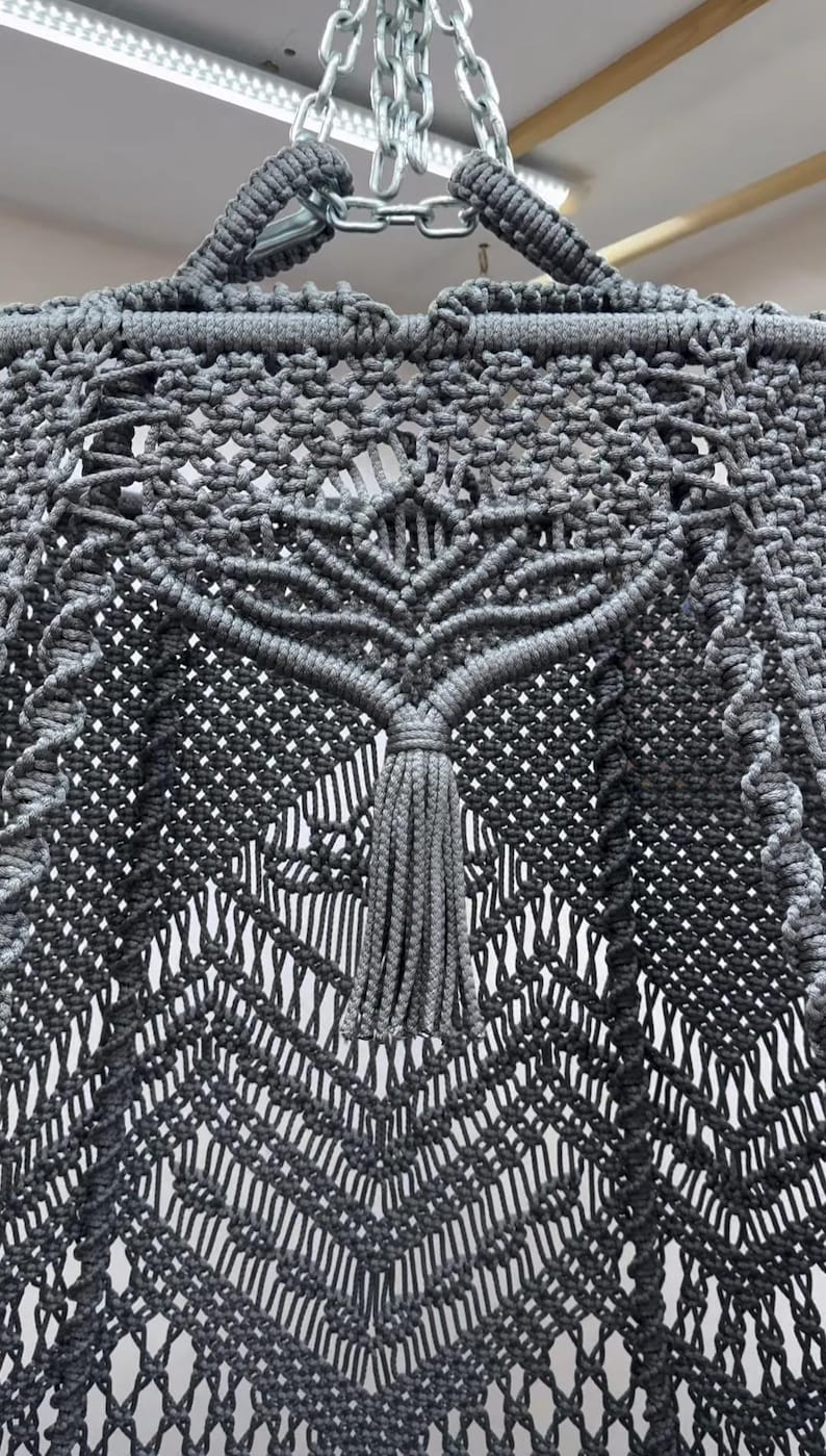 May include: Close-up of a gray macrame hammock chair. The chair features intricate knotting patterns, with a central design resembling a tree. A tassel hangs from the center. The chair is suspended by a metal chain.