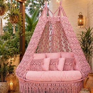 May include: A pink macrame hanging chair with a circular base and a canopy top. The chair features intricate knotted patterns, long fringe trim along the bottom, and includes several matching pink cushions arranged on the seat.