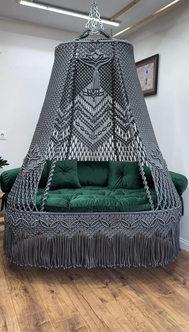 May include: A gray macrame hanging daybed with a canopy. The bed features a green velvet cushion and pillows. The macrame design includes intricate patterns and a fringe trim. The daybed is suspended from the ceiling, creating a cozy seating area.
