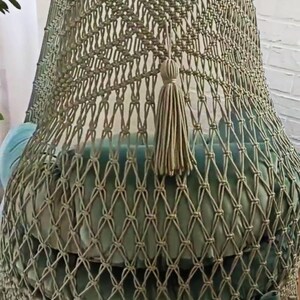 May include: A handcrafted macrame hanging chair in a soft sage green. The chair features a woven net design, a plush cushion, and a decorative tassel. The bottom edge is finished with fringe and teal ribbon ties, creating a bohemian aesthetic.