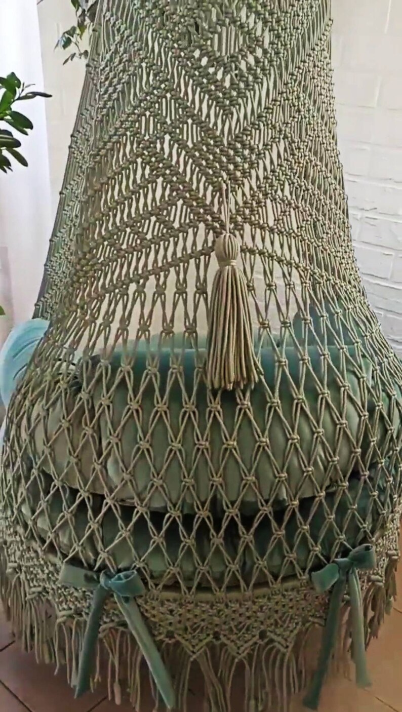 May include: A handcrafted macrame hanging chair in a soft sage green. The chair features a woven net design, a plush cushion, and a decorative tassel. The bottom edge is finished with fringe and teal ribbon ties, creating a bohemian aesthetic.