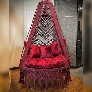 May include: A burgundy macrame hanging chair with a plush velvet cushion and fringe trim. The chair is suspended from the ceiling and has a bohemian style.