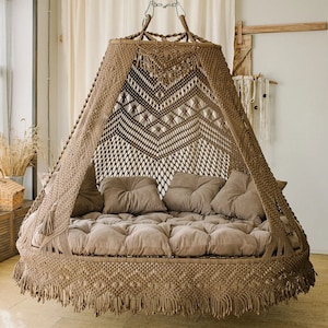 May include: A large, circular hanging swing chair featuring intricate tan macramé knotting in a geometric diamond pattern. The structure includes a canopy top, a tufted base cushion, and four matching square pillows in a solid light brown fabric.