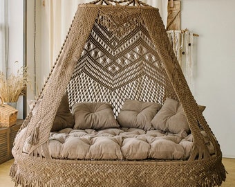 May include: A hanging macrame swing bed in a neutral brown color. The bed features a woven canopy with a geometric pattern, a cushioned base, and several throw pillows. The fringe detail adds a bohemian touch to this cozy seating area.