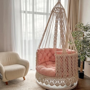May include: A pink and white macrame hanging chair with a pink cushion. The chair is suspended from the ceiling and is in a room with white curtains and a white rug.