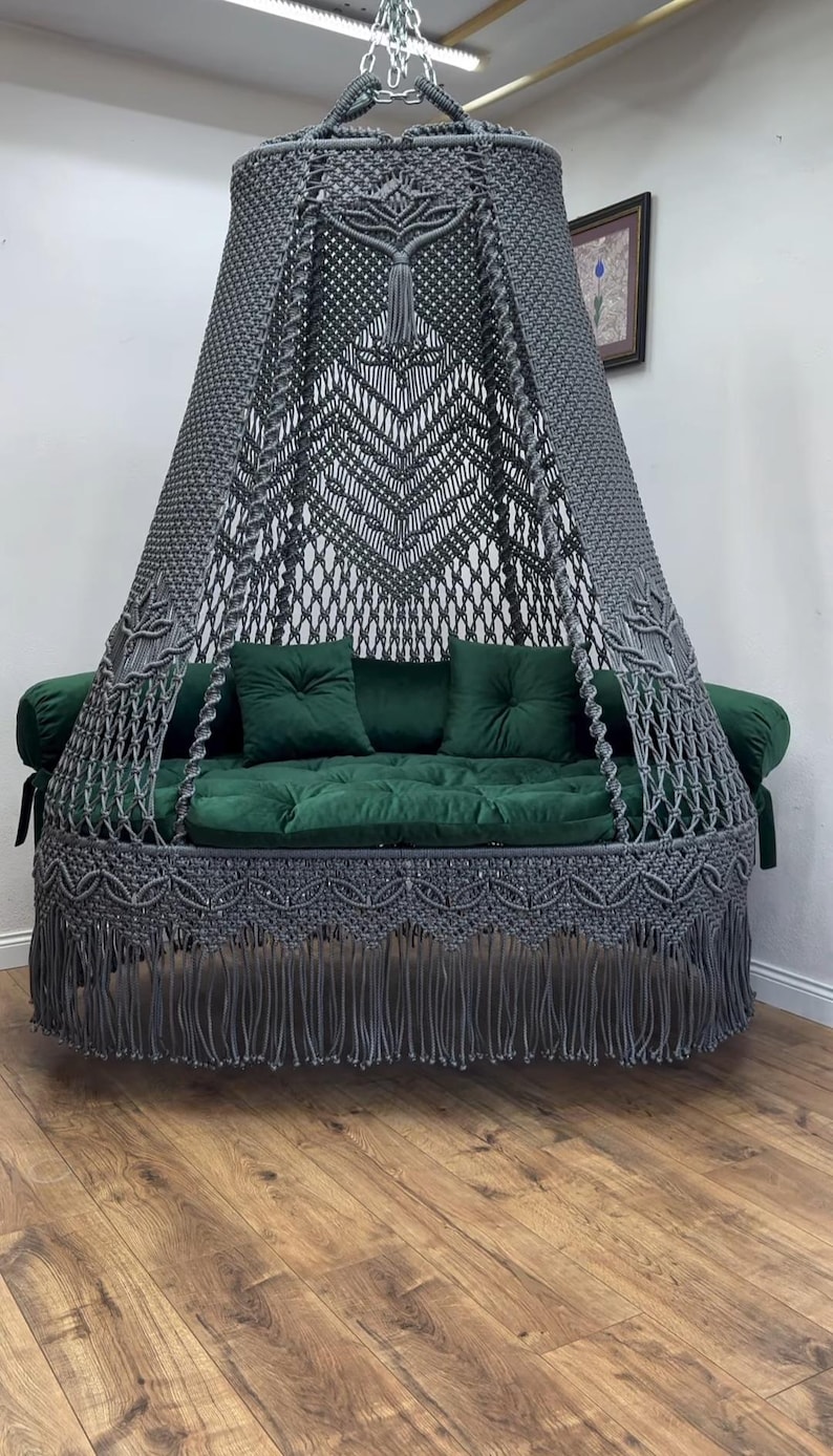 May include: A hanging macrame swing chair with a gray canopy and fringe. The seat features a green velvet cushion and two matching pillows. The chair is suspended from the ceiling and sits on a wooden floor.