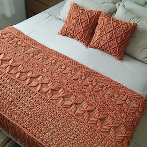 以下が含まれることがあります： 白い掛け布団のベッドには、織りの錆色のベッドランナーと、お揃いのクッションが2つ飾られています。 ベッドランナーは、下端にフリンジが付いた幾何学模様が特徴です。 クッションはダイヤモンドパターンです。