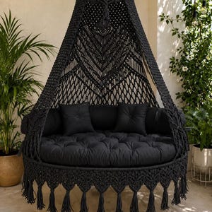 May include: A black macrame hanging chair featuring an intricate knotted canopy, a circular base with a thick tufted cushion, two matching square pillows, and a decorative fringe border with long tassels suspended from a central metal hook.