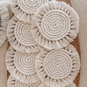 May include: Set of six round crocheted coasters with fringe. The coasters are made of natural cream-colored cotton yarn and have a spiral design.