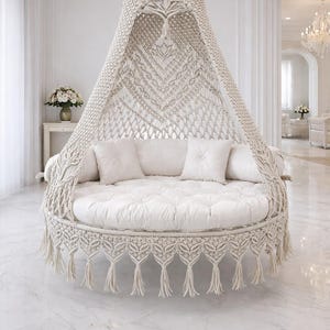 May include: A cream-colored macrame hanging daybed with a round, cushioned base and a canopy. The bed features a fringe trim and is adorned with white pillows. It is suspended from the ceiling in a bright, white room with marble flooring.