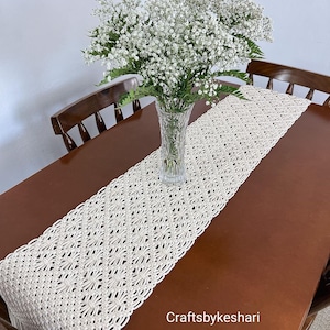 May include: A white crocheted table runner with a diamond pattern and fringe edges. The runner is on a wooden table with a vase of white baby's breath flowers. The text "Craftsbykeshari" is visible in the lower right corner of the image.