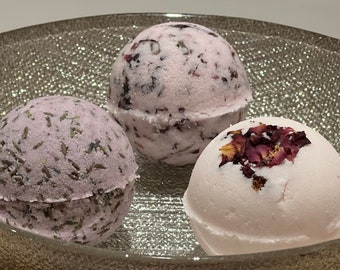 Op de afbeelding: Drie badbommen in een zilveren kom. De badbommen zijn roze en wit met daarin gedroogde bloemen en kruiden.