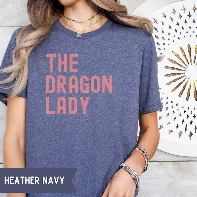 Pode incluir: T-shirt azul marinho mesclado com o texto "THE DRAGON LADY" em letras coral. A camisa &eacute; de gola redonda e tem mangas curtas. As palavras "HEATHER NAVY" s&atilde;o impressas na parte inferior da imagem.