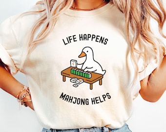May include: Cream-colored sweatshirt with the text "LIFE HAPPENS MAHJONG HELPS". The graphic features a cartoon duck playing mahjong. The duck is sitting at a brown table with green tiles and white tiles.