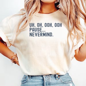 May include: A cream-colored short-sleeve t-shirt featuring dark blue text printed on the chest that reads, UH, OH, OOH, OOH PAUSE... NEVERMIND. The shirt has a crew neckline and a relaxed fit, paired with blue denim jeans visible at the waistline.