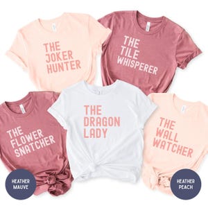 May include: Five short-sleeved t-shirts in various colors, each with a different phrase printed in pink. The phrases include "The Joker Hunter," "The Tile Whisperer," "The Flower Snatcher," "The Dragon Lady," and "The Wall Watcher."