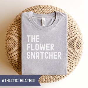 Pode incluir: Camiseta cinza mesclada com a frase branca "THE FLOWER SNATCHER". A camiseta est&aacute; dobrada e colocada sobre uma superf&iacute;cie circular tecida. Uma pequena etiqueta branca &eacute; vis&iacute;vel perto da gola.